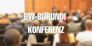 Man sieht viele Menschen von hinten, die in Stuhlreihen sitzen. Ganz vorne steht auf der Bühne ein Redner. Auf dem Bild steht BW-Burundi Konferenz.